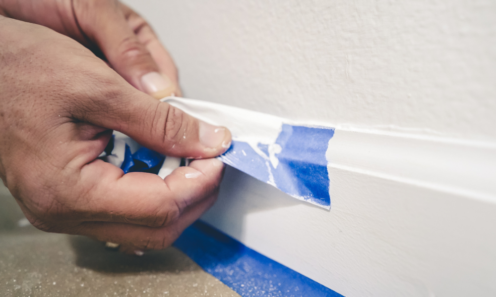 A painter pulls of blue painter's tape from the wall to reveal a clean edge baseboard.